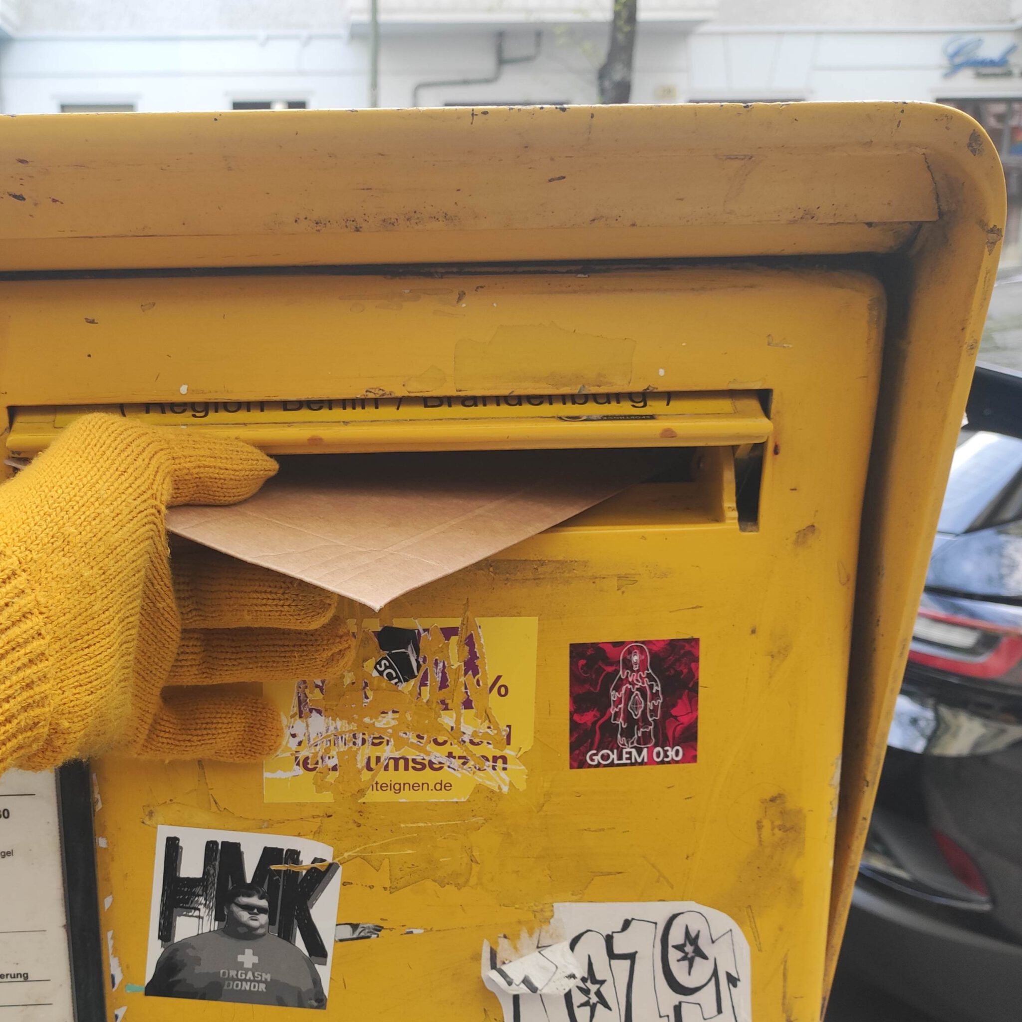 Eine Hand mit gelbem Wollhandschuh wirft einen braunen Umschlag in einen gelben Briefkasten
