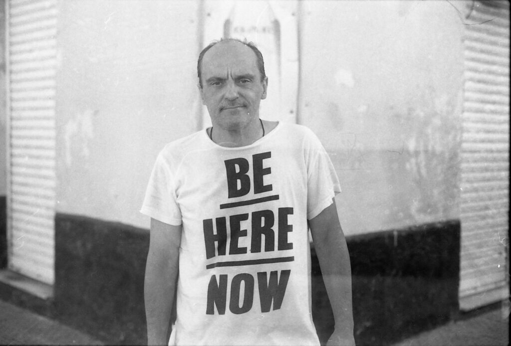 Schwarz-weiß Foto eine Mannes mit Halbglatze mit Tshirt mit der Aufschrift Be Here Now